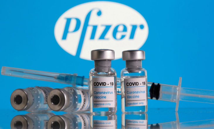 新型コロナワクチンのラベルが貼られた薬剤と注射器。後ろはファイザー社のロゴ （Dado Ruvic/Reuters）